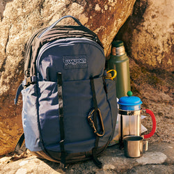 Blue Jansport backpack with a thermos and coffee press against a rock background