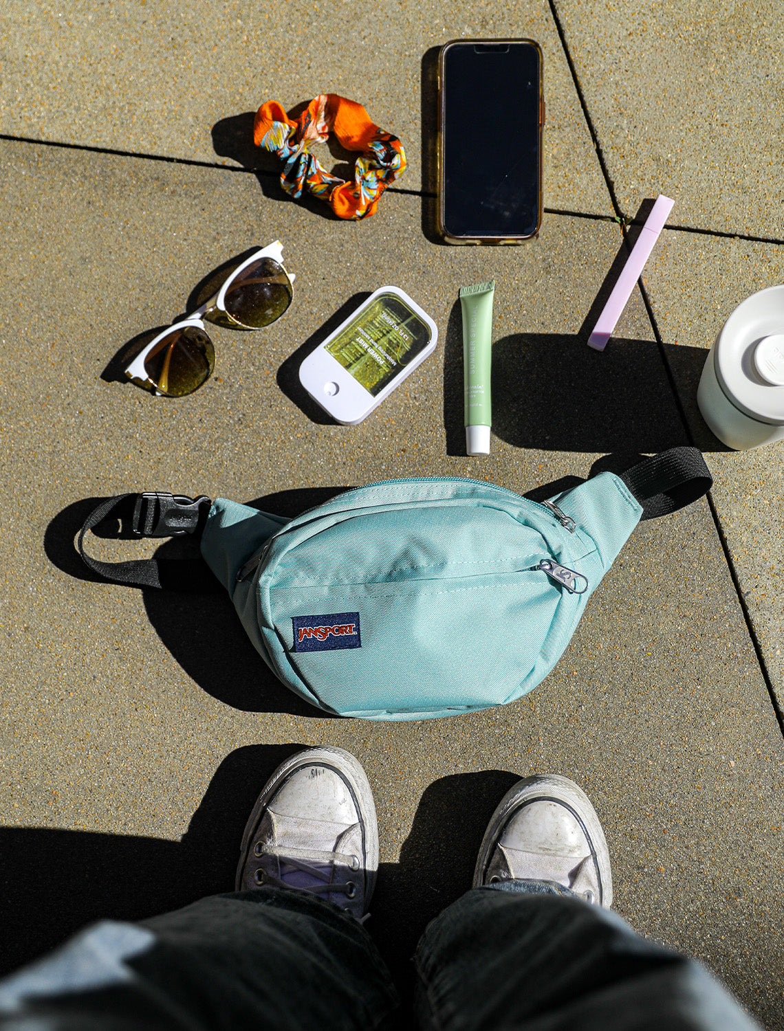 Light blue JanSport waist bag on a concrete surface with sunglasses, phone, scrunchie, and other items.