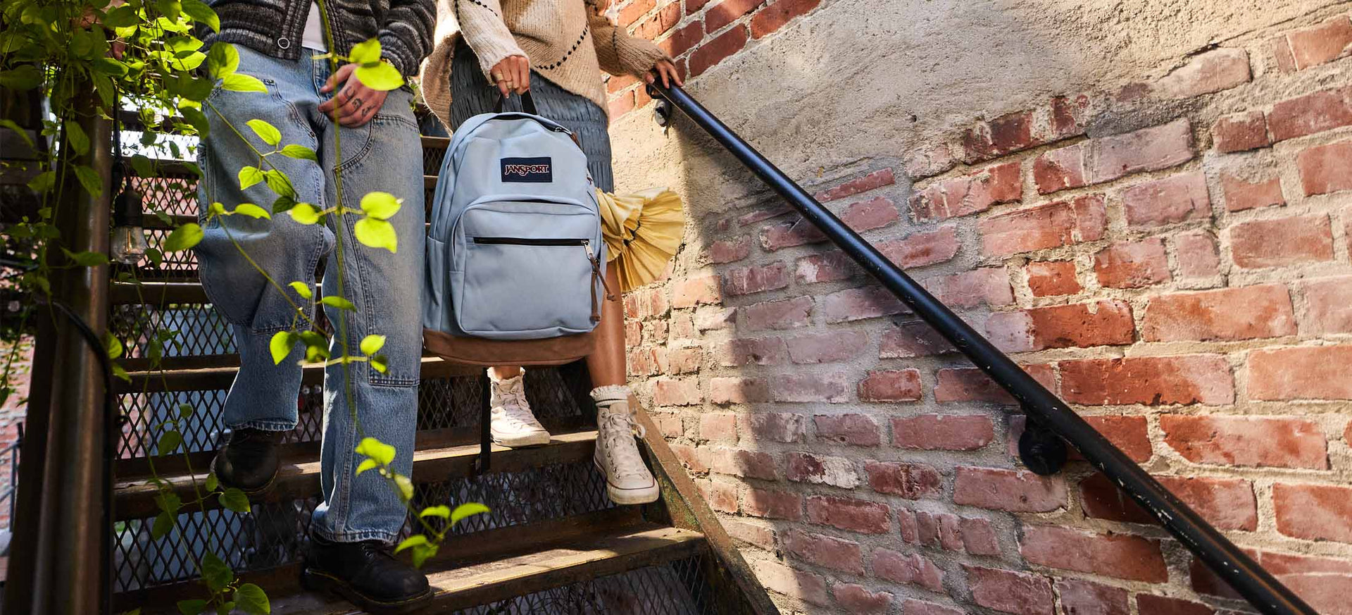 Two people walking up a set of stairs holding a jansport light blue right pack with a brick wall in the background